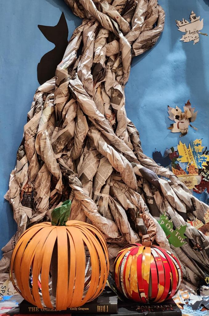 Cardboard pumpkins setting on book-cover paper leaves in front of a haunted tree made of newspaper. The background is blue.